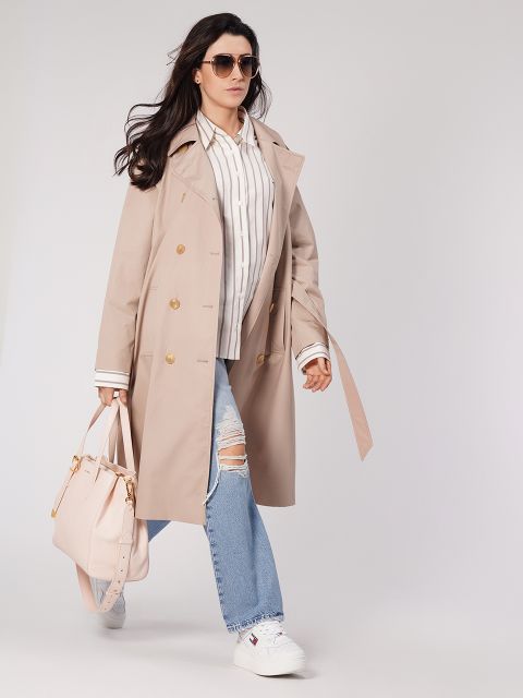 Eine Person mit langen Haaren, Sonnenbrille, beigem Trenchcoat, gestreiftem Hemd, zerrissenen Jeans und weißen Turnschuhen trägt eine helle Handtasche. Sie läuft selbstbewusst vor einem neutralen Hintergrund.