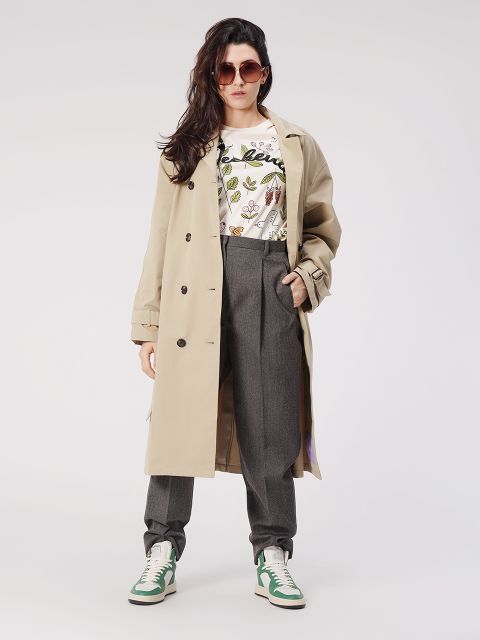 Eine Person in beigem Trenchcoat, gemustertem Hemd, grauer Hose, grün-weißen Sneakers und Sonnenbrille posiert selbstbewusst vor einem schlichten Hintergrund. Ihr Haar ist lang und in sanften Wellen gestylt.