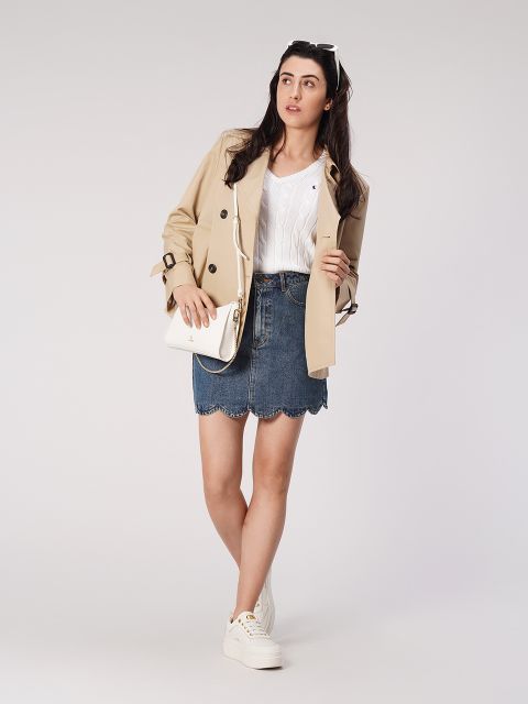 Eine Frau steht vor einem schlichten Hintergrund. Sie trägt einen beigen Trenchcoat, ein weißes Oberteil und einen Jeansrock. Sie hat eine kleine weiße Umhängetasche und weiße Turnschuhe dabei. Ihre langen Haare sind offen und sie trägt eine Sonnenbrille auf dem Kopf.