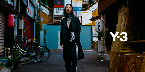 Eine Person in einem schwarzen Y-3-Outfit mit weißen Grafiken steht in einer städtischen Gasse. Fahrräder und verschiedene Ladenschilder säumen die Straßenränder.