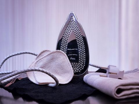 Ein Bügeleisen steht aufrecht auf einer reflektierenden Oberfläche, umgeben von einem glatt gebügelten Hemd, einer Stoffmaske und einer Kordel. Der Hintergrund ist verschwommen und hebt die häusliche Umgebung hervor.