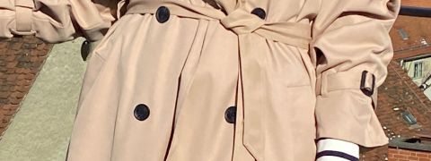 Eine Person trägt einen beigefarbenen zweireihigen Trenchcoat mit Gürtel in der Taille und steht vor einer Dächerkulisse. Der Mantel hat dunkle Knöpfe und verstellbare Manschetten.