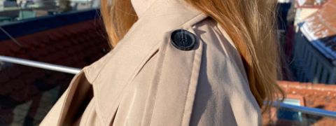 Eine Person mit langen Haaren trägt einen beigen Trenchcoat auf einem Dach. Der Fokus liegt auf der Schulter des Mantels mit einem dekorativen Knopf. Im Hintergrund sind Dächer und ein klarer Himmel zu sehen.