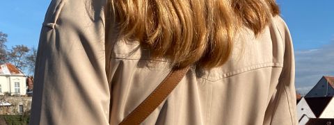 Eine Person mit langem, hellbraunem Haar, das den Blick abwendet, trägt ein beiges Hemd und einen braunen Schultergurt. Sie ist im Freien mit einem klaren blauen Himmel und teilweiser Sicht auf Gebäude und Bäume im Hintergrund.