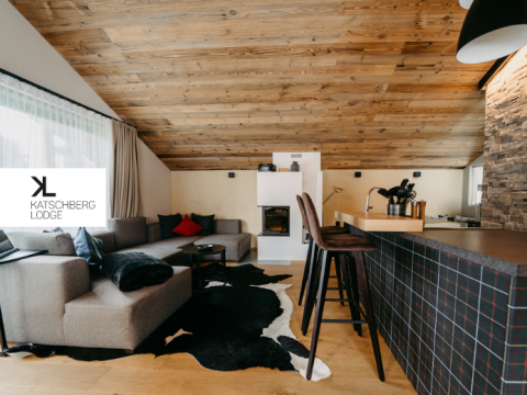 Ein gemütlicher moderner Wohnbereich mit Holzdecke, einem Sofagarnitur, einem Kuhfellteppich, einem Kamin und einer Kücheninsel mit Barhockern. In der Ecke prangt ein Logo mit der Aufschrift „Katschberg Lodge“.