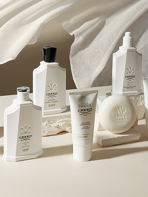 Eine luxuriöse Präsentation von Creed-Hautpflege- und Duftprodukten, darunter Flaschen, eine Tube und ein Stück Seife. Alle Artikel sind elegant auf einer neutral getönten Oberfläche mit weichem, fließendem Stoff im Hintergrund angeordnet.
