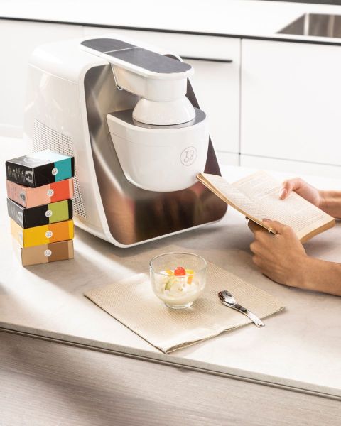 Eine Person liest ein Buch neben einer Eismaschine auf einer Küchentheke. Daneben steht eine Schüssel Eiscreme mit einer Kirsche darauf, ein Löffel und ein Stapel bunter Schachteln.