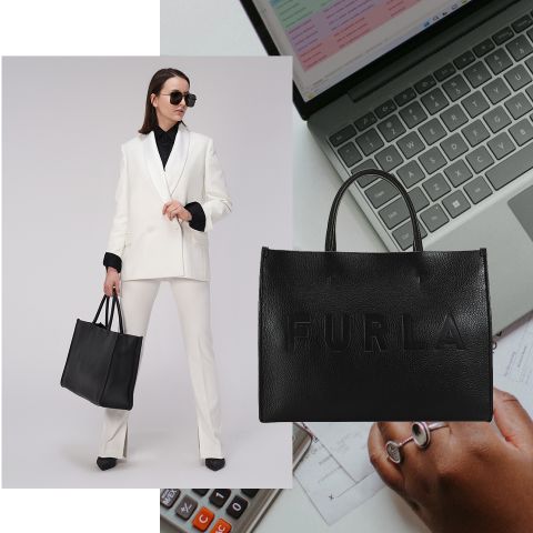 Eine Frau in einem weißen Anzug und einer schwarzen Bluse hält eine schwarze Furla-Handtasche. Sie trägt eine Sonnenbrille und steht selbstbewusst da. Im Hintergrund sind ein Laptop, ein Taschenrechner und eine beringte Hand zu sehen.