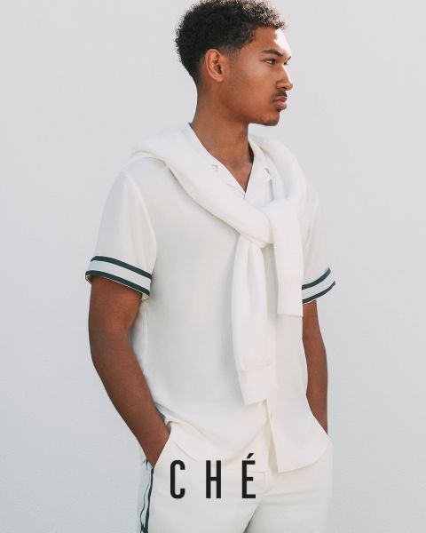 Ein Mann in weißem Outfit mit schwarzem Besatz, bestehend aus kurzärmeligem Hemd und Hose, posiert vor einem schlichten weißen Hintergrund. Ein weißer Pullover hängt über seinen Schultern, die Hände stecken in seinen Taschen. Unten steht der Text „CHÉ“.