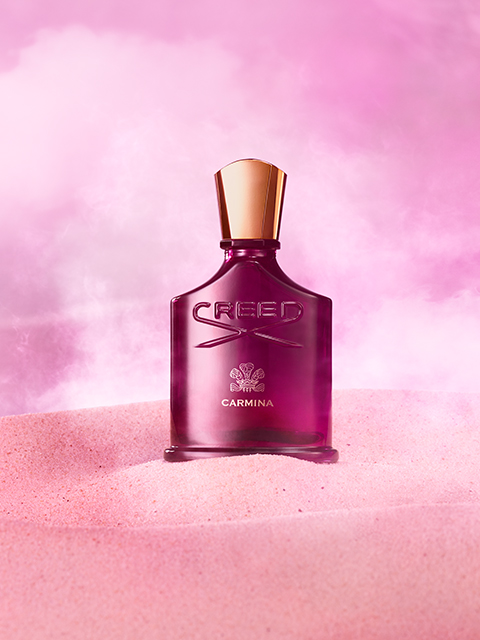 Eine rosa Flasche Creed Carmina-Parfüm steht vor einem rosa nebligen Hintergrund und ruht auf einer Oberfläche, die rosa Sand ähnelt. Die Flasche hat einen goldenen Verschluss und zeigt den Markennamen und das Logo in erhabener Schrift.