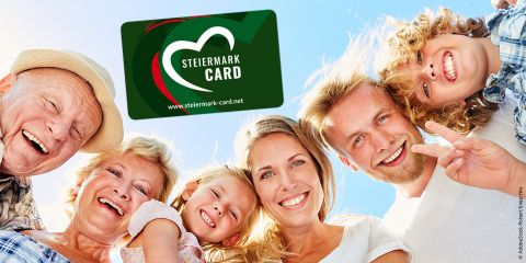 Eine Gruppe von sechs lächelnden Menschen, darunter Erwachsene und Kinder, drängt sich unter einem klaren blauen Himmel zusammen. Über ihnen schwebt eine Steiermark Card mit einem Herzlogo und einem Website-Link.