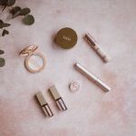 Verschiedene Make-up-Produkte, angeordnet auf einer strukturierten rosa Oberfläche, darunter Lidschatten, Lippenstifte und ein kleiner Pflanzenzweig auf der linken Seite.