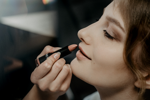 Nahaufnahme einer Person mit braunen Augen, die leicht lächelt, während Lippenstift auf ihre Lippen aufgetragen wird. Der Lippenstift hat einen sanften, neutralen Farbton und die Person trägt dezentes Augen-Make-up. Der Hintergrund ist unscharf.