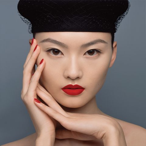 Eine Person mit auffällig rotem Lippenstift und Nagellack posiert vor einem grauen Hintergrund. Sie trägt einen schwarzen Hut mit Netzstruktur und ihre Hände sind elegant um ihr Gesicht gelegt, was das leuchtende Make-up betont.