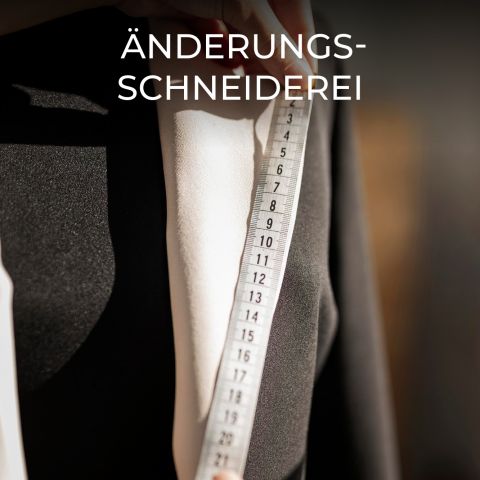 Nahaufnahme einer Person, die ein Maßband an ein weißes Hemd unter einem schwarzen Blazer hält, mit dem Text „Änderungs-Schneiderei“ oben drauf. Die Szene lässt auf Schneiderei oder Änderungen schließen.