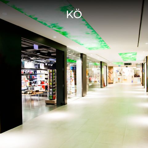 Ein moderner Korridor eines Einkaufszentrums mit hell erleuchteten Geschäften auf beiden Seiten. Die Decke ist mit grünen abstrakten Mustern verziert. Oben im Bild ist das Wort „KÖ“ zu sehen.