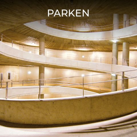 Eine mehrstöckige spiralförmige Parkrampe mit Betonwänden und Metallgeländern, beleuchtet von hellen Lichtern. Oben im Bild ist das Wort „PARKEN“ zu sehen. Die Architektur zeichnet sich durch zylindrische Säulen und geschwungene Wege aus.