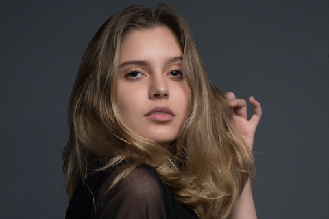 Eine Person mit langen, welligen blonden Haaren posiert vor einem dunkelgrauen Hintergrund. Sie trägt ein schwarzes Oberteil und hat einen ruhigen Gesichtsausdruck, wobei sie mit einer Hand ihr Haar berührt.
