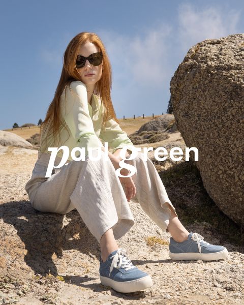 Eine Person mit langen roten Haaren, einer Sonnenbrille, einem hellgrünen Oberteil, beigen Hosen und blauen Turnschuhen sitzt auf felsigem Gelände unter blauem Himmel. Die Worte „Paul Green“ sind über das Bild gelegt.