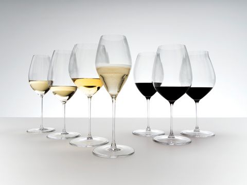 Ein Arrangement aus sieben Weingläsern auf einer weißen Oberfläche, jedes gefüllt mit unterschiedlichen Weinsorten, von Weiß- bis Rotwein. Die Gläser sind elegant geformt und leicht geneigt, wodurch die Vielfalt an Farbe und Transparenz zur Geltung kommt.