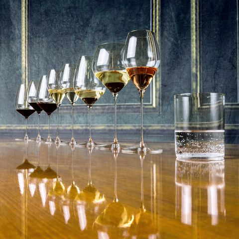 Eine Reihe von sieben Weingläsern, jedes mit unterschiedlichen Weinsorten und -mengen gefüllt, steht auf einer reflektierenden Holzoberfläche. Daneben steht auch ein Glas mit klarem Sprudelwasser. Der Hintergrund besteht aus strukturierten grünen Wänden mit goldenen Akzenten.