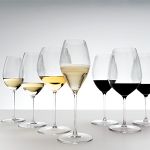 Sieben in einer Reihe angeordnete Weingläser, gefüllt mit verschiedenen Weinsorten. Die Gläser auf der linken Seite enthalten Weißwein, das mittlere Glas enthält Sekt und die rechten Gläser sind mit Rotwein gefüllt, vor einem Hintergrund mit Farbverlauf.