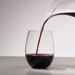 Rotwein wird aus einer Karaffe in ein klares, stielloses Glas auf einer glatten, reflektierenden Oberfläche vor einem minimalistischen, grauen Hintergrund gegossen.