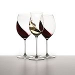 Drei Weingläser mit wirbelndem Wein darin. Zwei Gläser enthalten Rotwein und eines Weißwein. Die Gläser sind elegant auf einer glatten Oberfläche vor einem schlichten weißen Hintergrund angeordnet.