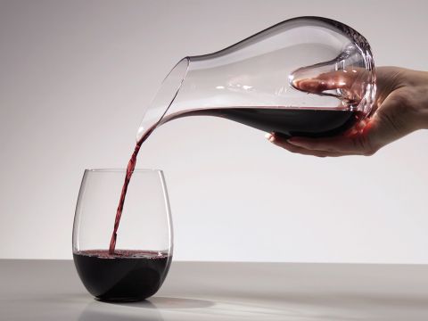 Eine Hand gießt Rotwein aus einer durchsichtigen Glaskaraffe in ein stielloses Weinglas auf einer hellgrauen Oberfläche. Der Hintergrund ist sanft beleuchtet, was den dunklen Farbton des Weins und die eleganten Kurven der Karaffe betont.