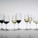 Eine Vielzahl von Weingläsern, jedes mit unterschiedlichen Weinsorten gefüllt, sind in einer Reihe auf einer reflektierenden Oberfläche angeordnet. Die Sammlung umfasst Rot- und Weißweine sowie Champagner vor einem einfachen weißen Hintergrund.