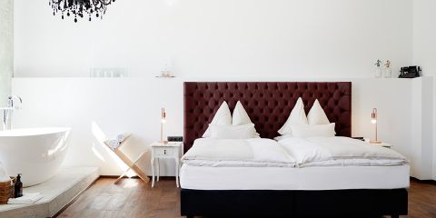 Modernes Hotelzimmer mit großem Bett und dunklem gepolstertem Kopfteil. Weiße Bettwäsche bildet einen Kontrast zum dunklen Holzboden. Links steht eine freistehende Badewanne, zusammen mit einem kleinen Tisch und Stuhl. Darüber hängt ein schwarzer Kronleuchter, der Eleganz verleiht. Minimalistisches Dekor.