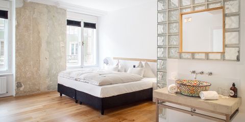 Ein modernes Schlafzimmer mit einem großen Bett mit weißer Bettwäsche neben einer strukturierten Wand. Der Raum hat Holzfußboden, ein Fenster mit schwarzen Jalousien und einen offenen Badezimmerbereich mit einem farbenfrohen, gemusterten Waschbecken und einer Trennwand aus Glasbausteinen.