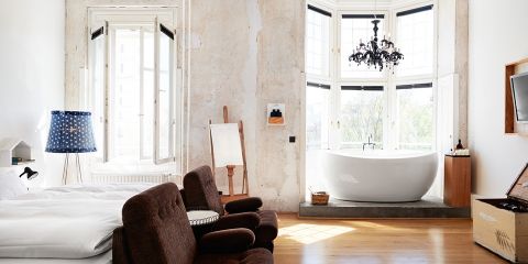 Ein stilvolles Schlafzimmer mit offenem Design. Es verfügt über ein großes Bett, braune Stühle, eine freistehende Badewanne neben einem Erkerfenster, eine Staffelei und einen Kronleuchter. Licht fällt durch die Fenster und unterstreicht die Mischung aus rustikalem und modernem Dekor.