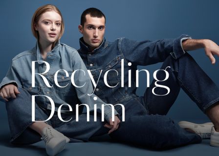 Zwei Models posieren vor einem blauen Hintergrund und tragen Jeans in verschiedenen Farbtönen. Der Text auf dem Bild lautet „Recycling Denim“. Das männliche Model hat kurze Haare und das weibliche Model hat lange Haare. Beide sitzen lässig da.