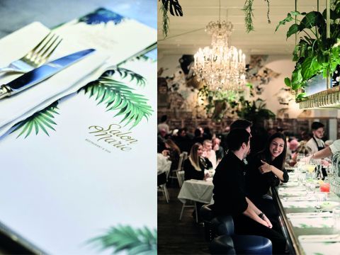 Eine stilvolle Restaurantszene: Links eine Speisekarte und Besteck auf einem Tisch mit Blättermuster. Rechts ein lebhafter Essbereich mit Leuten an einer Bar, kunstvollen Kronleuchtern und Hängepflanzen, die die Atmosphäre bereichern.