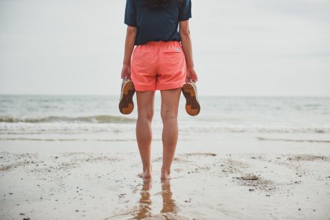 Eine Person steht barfuß an einem Sandstrand mit Blick auf das Meer und hält in jeder Hand Sandalen. Sie trägt korallenfarbene Shorts und ein dunkles Oberteil. Der Himmel ist bedeckt und sanfte Wellen nähern sich dem Ufer.