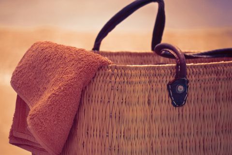 Eine Strandtasche aus Korbgeflecht mit dunklen Griffen liegt vor einem sandigen Hintergrund. Über den Rand der Tasche hängt ein gefaltetes orangefarbenes Handtuch, das an einen sonnigen Tag am Strand erinnert.