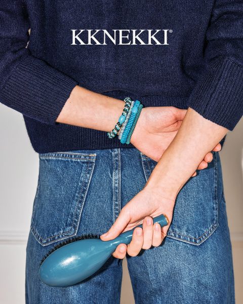 Eine Person in einem marineblauen Pullover und Jeans hält eine blaue Haarbürste hinter ihrem Rücken und trägt türkisfarbene Perlenarmbänder. Darüber das Logo "KKNEKKI".