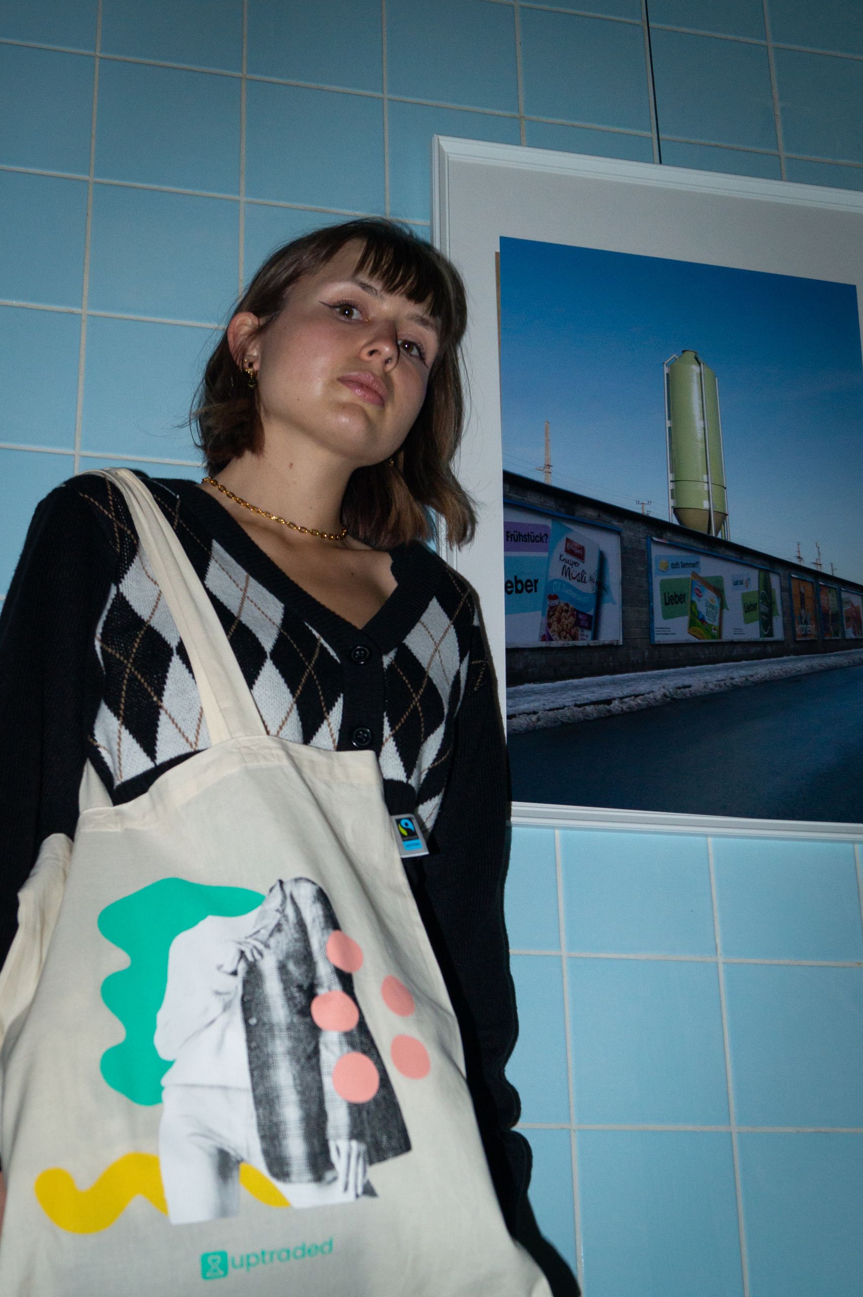 Eine Person mit kurzen Haaren steht vor einer blau gefliesten Wand und trägt einen schwarz-weißen Pullover. Über einer Schulter trägt sie eine Einkaufstasche mit abstrakter Kunst. An der Wand neben ihr hängt ein gerahmtes Foto eines Gebäudes.