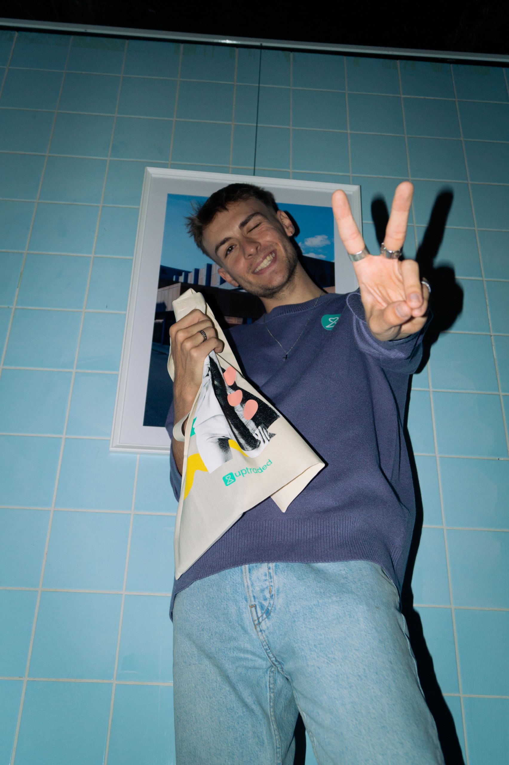 Eine lächelnde Person hält eine Einkaufstasche und zeigt mit der Hand das Peace-Zeichen. Sie trägt einen lila Pullover und helle Jeans und steht vor einer blau gefliesten Wand mit einem gerahmten Bild.