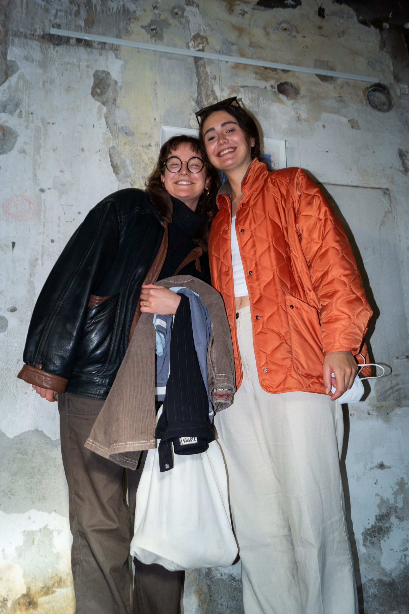 Zwei lächelnde Menschen stehen vor einer verwitterten Wand. Einer trägt eine Brille, eine dunkle Jacke und hält einen Mantel, während der andere eine orangefarbene Jacke trägt und eine Brille hält. Beide wirken entspannt und glücklich.