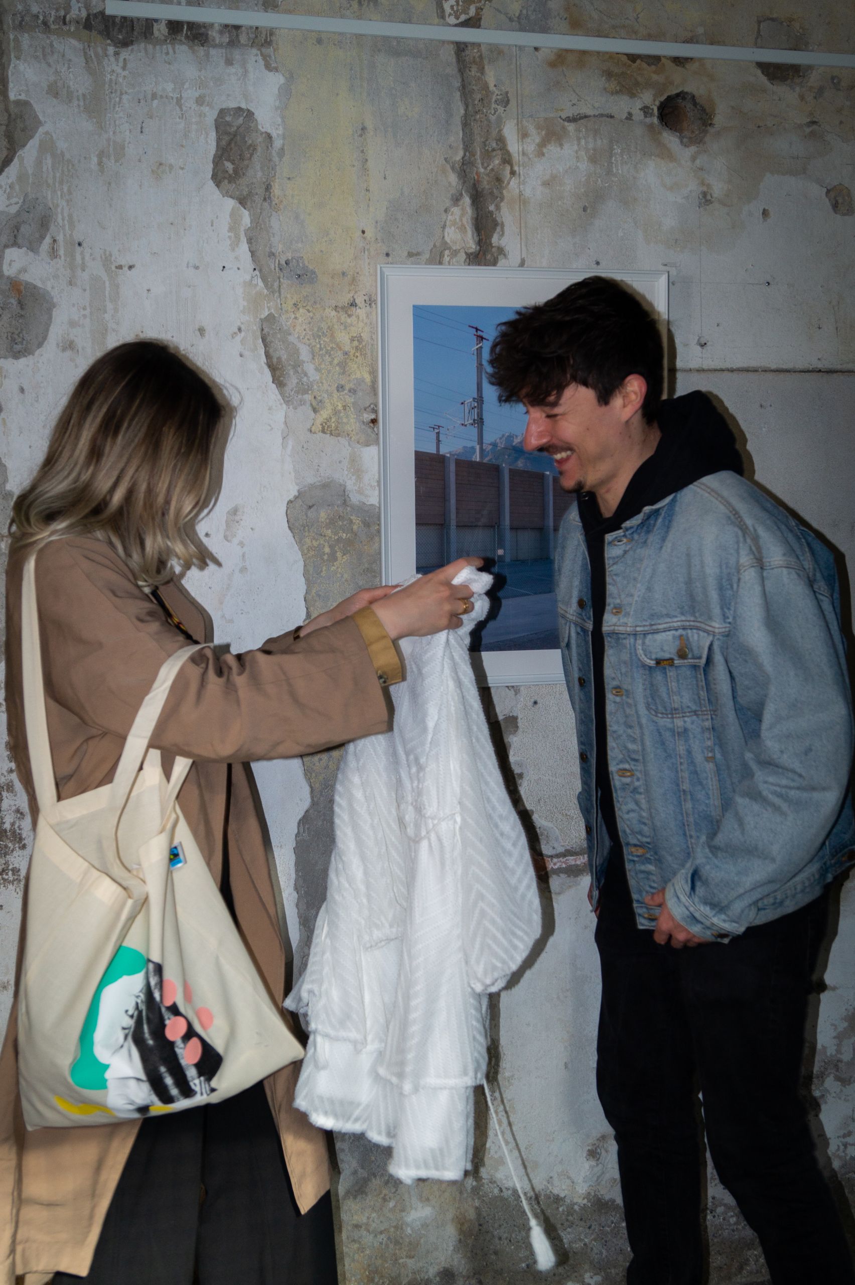 Eine Frau mit Einkaufstasche zeigt einem Mann in Jeansjacke ein weißes Kleidungsstück. Sie stehen vor einer verwitterten Wand mit einem kleinen gerahmten Bild. Beide wirken amüsiert oder in ein Gespräch vertieft.