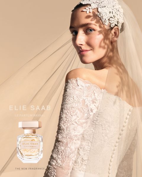 Braut in Spitzenkleid und Schleier, mit Blumenhaarteil, blickt über ihre Schulter. Ihr Kleid wirft einen weichen Schatten mit Blumenmuster auf beigem Hintergrund. Ein Parfümflakon mit dem Text „Elie Saab Le Parfum Bridal“ ist ausgestellt.