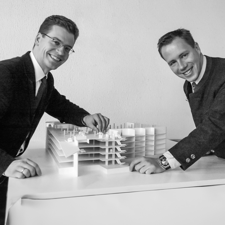 Zwei Männer in Anzügen lächeln und beugen sich über einen weißen Tisch mit einem detaillierten Architekturmodell einer mehrschichtigen Struktur. Sie scheinen ihr Projekt in einem professionellen Umfeld zu diskutieren oder vorzustellen.