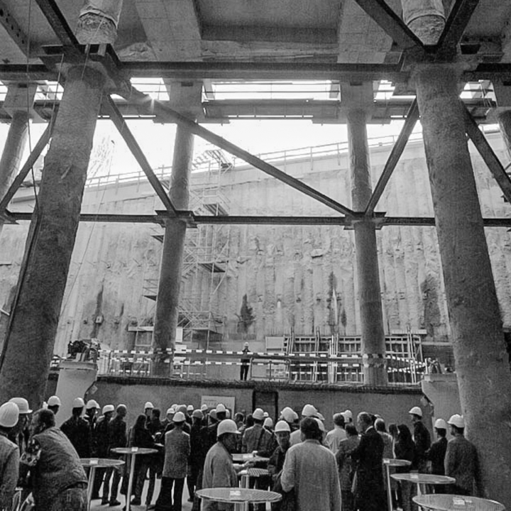 Eine Gruppe von Menschen mit Schutzhelmen steht auf einer großen Baustelle mit hoch aufragenden Betonpfeilern und einer hohen Mauer im Hintergrund. Rund um die Baustelle sind Gerüste und Baumaterialien zu sehen.