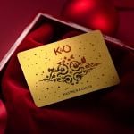 Eine goldene Geschenkkarte mit kunstvollen schwarzen Motiven und dem Text „K&Ö“ und „Kastner & Öhler“ liegt auf einem satten roten Stoff und ist in einer offenen roten Schachtel untergebracht. Der Hintergrund ist mit roten Weihnachtsornamenten verziert, die für eine festliche Atmosphäre sorgen.