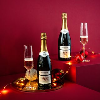 Zwei Champagnerflaschen mit gefüllten Gläsern, platziert auf einem roten Hintergrund mit festlichen Lichterketten und Ornamenten. Die Szene ist auf erhöhten Plattformen aufgebaut und schafft eine festliche und elegante Atmosphäre.