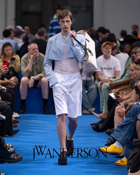 Ein Model auf einem blauen Laufsteg trägt ein hellblaues kurzes Shirt, weiße Shorts und schwarze Plateauschuhe und trägt eine große weiße Tasche. Sitzende Zuschauer schauen zu und machen Fotos. Das Logo von J.W. Anderson ist auf dem Boden zu sehen.
