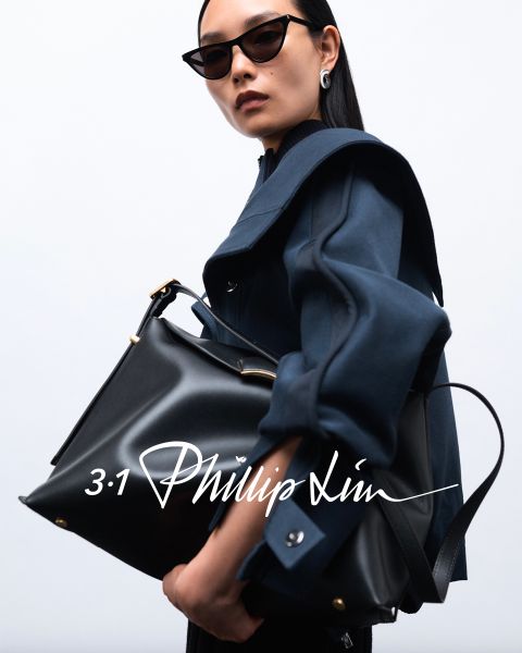 Person mit Sonnenbrille, dunklem Mantel und einer großen schwarzen Tasche vor einem schlichten Hintergrund. Auf der Textüberlagerung steht in stilisierter Handschrift „3.1 Phillip Lim“.
