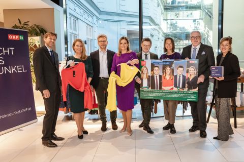Eine Gruppe von acht Personen steht zusammen in einem Innenbereich und lächelt in die Kamera. Zwei halten bunte Kleidungsstücke hoch, während andere ein großes Werbeplakat und eine Broschüre halten. Sie scheinen Teil einer Veranstaltung oder Kampagne zu sein. Auf einem Schild links steht „LICHT INS DUNKEL“.